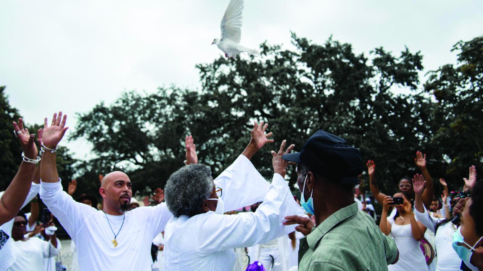 A group of Black people dressed in white release doves into the air in an outside environment.