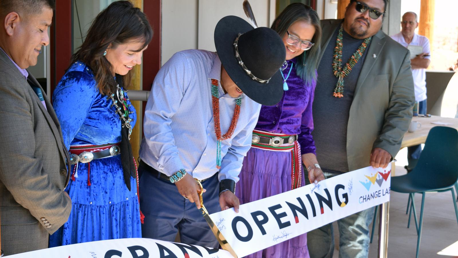 A small group of people stand behind a banner reading "Grand Opening". A man in the center is cutting the banner with large golden scissors.