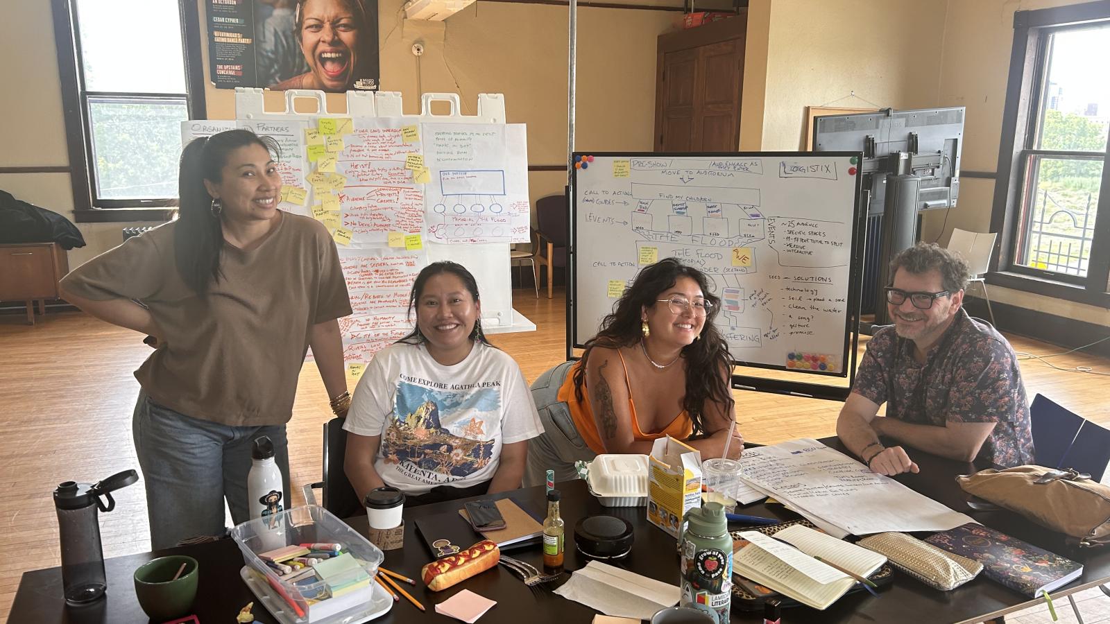 A man and three women are seated at or stand behind a table, smiling at the camera. The table is strewn with papers and writing utensils, and behind them are large boards covered in writing and post-its.
