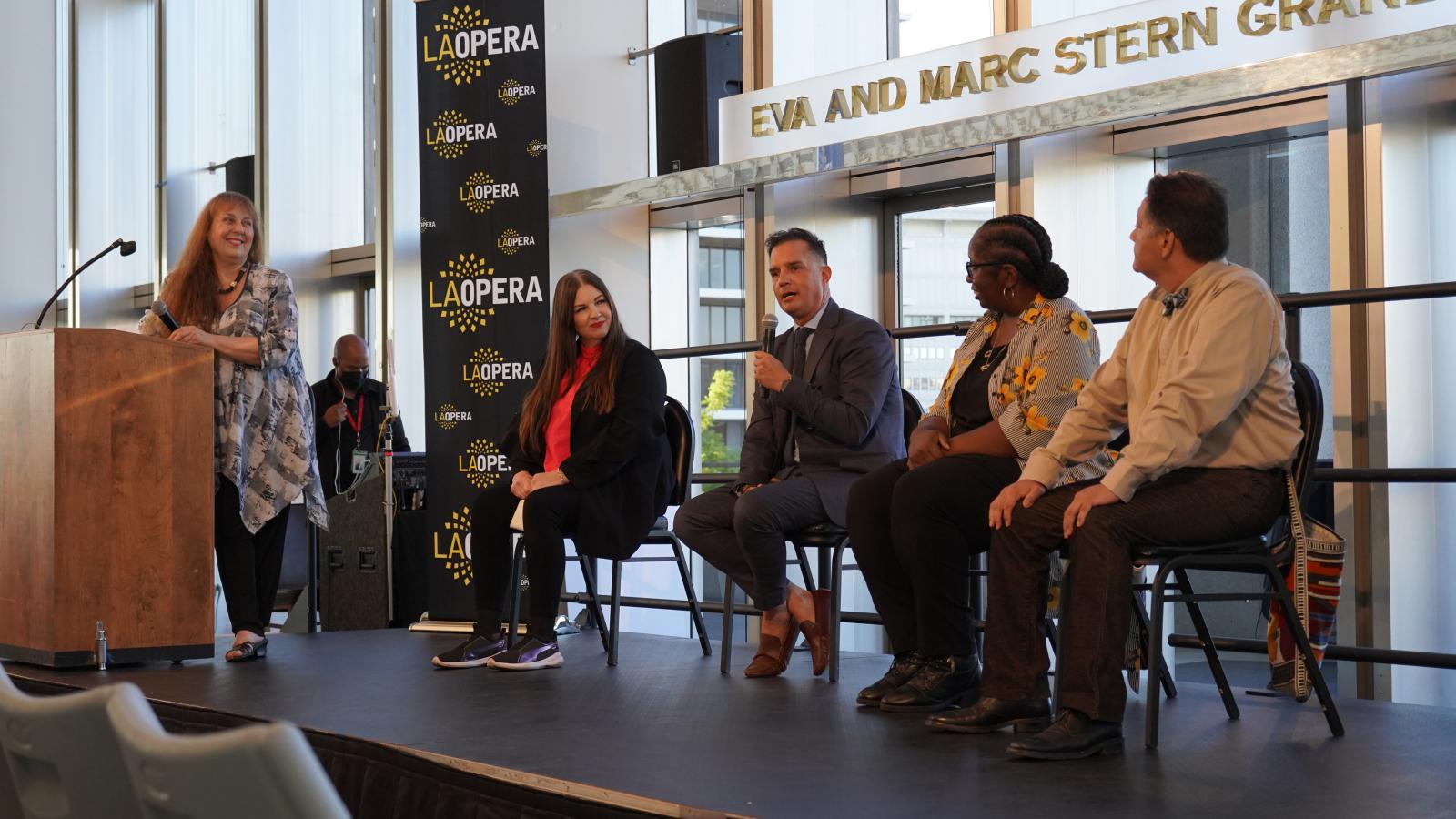 Four people seated on chairs on stage, man second from left holding microphone, woman standing at lectern to the left.
