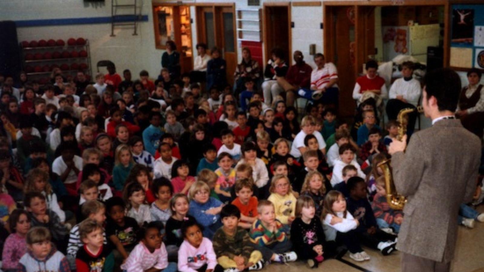 A large group of young children sitting cross-legged on the floor listening to a man play a saxophone