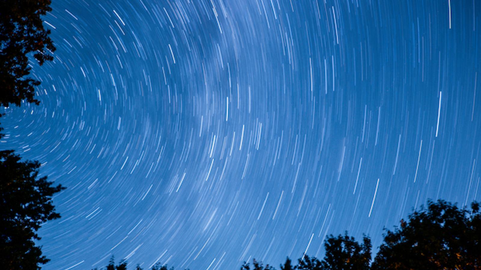 arcs of light in a darkening sky with a line of trees at bottom of frame