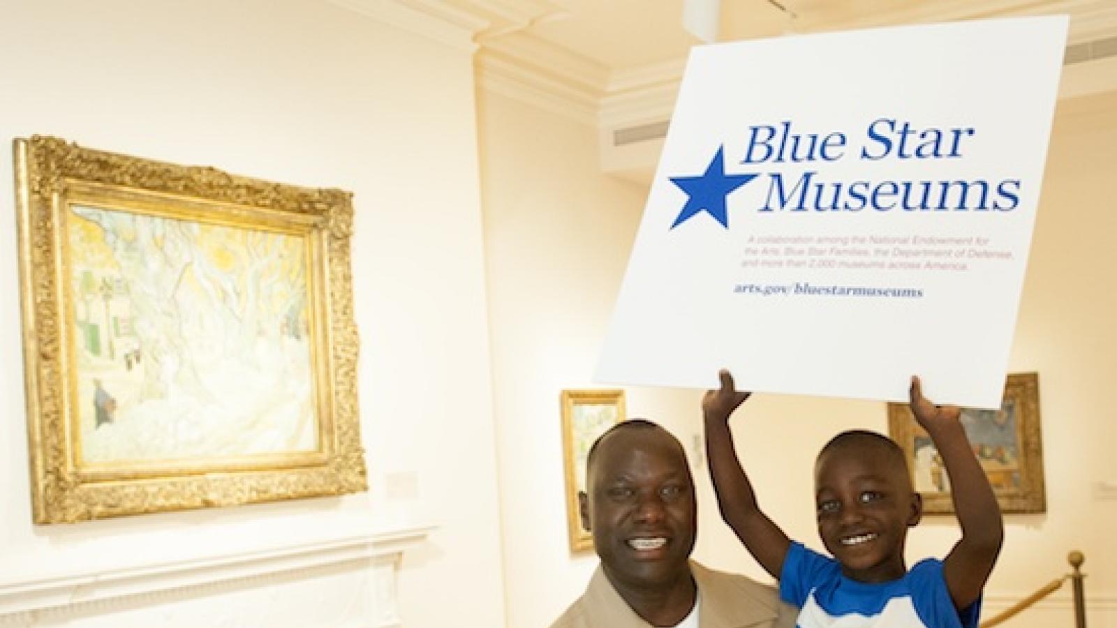An African-American man in a Marine uniform holds his son in an art museum with artworks in the background