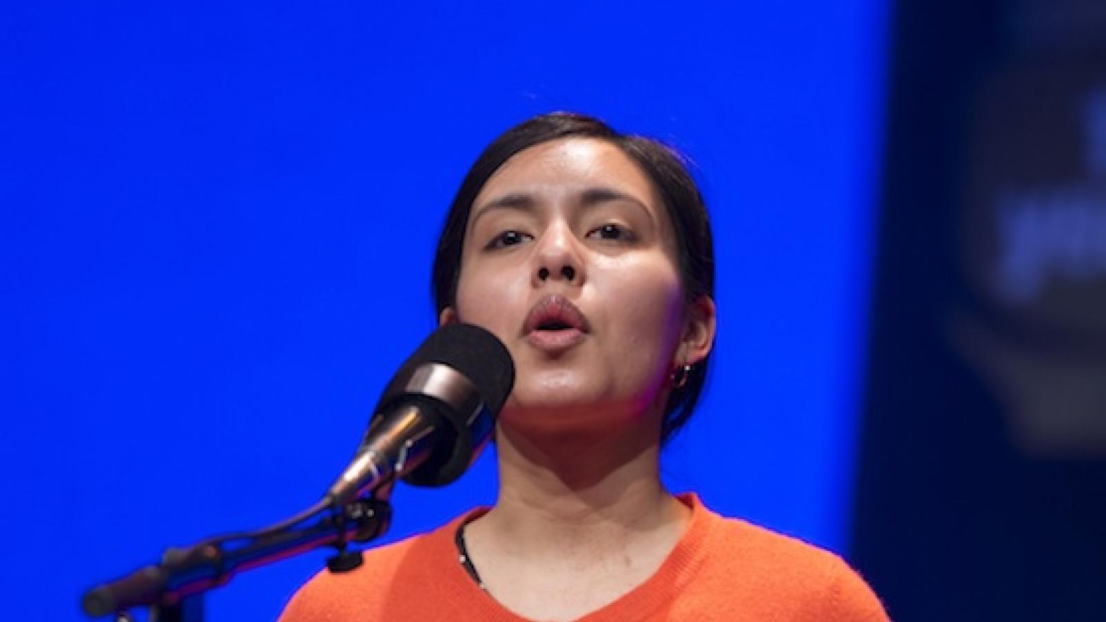 a young Hispanic woman wearing an unbuttoned orange sweater over a black dress with white polka dots recites into a microphone