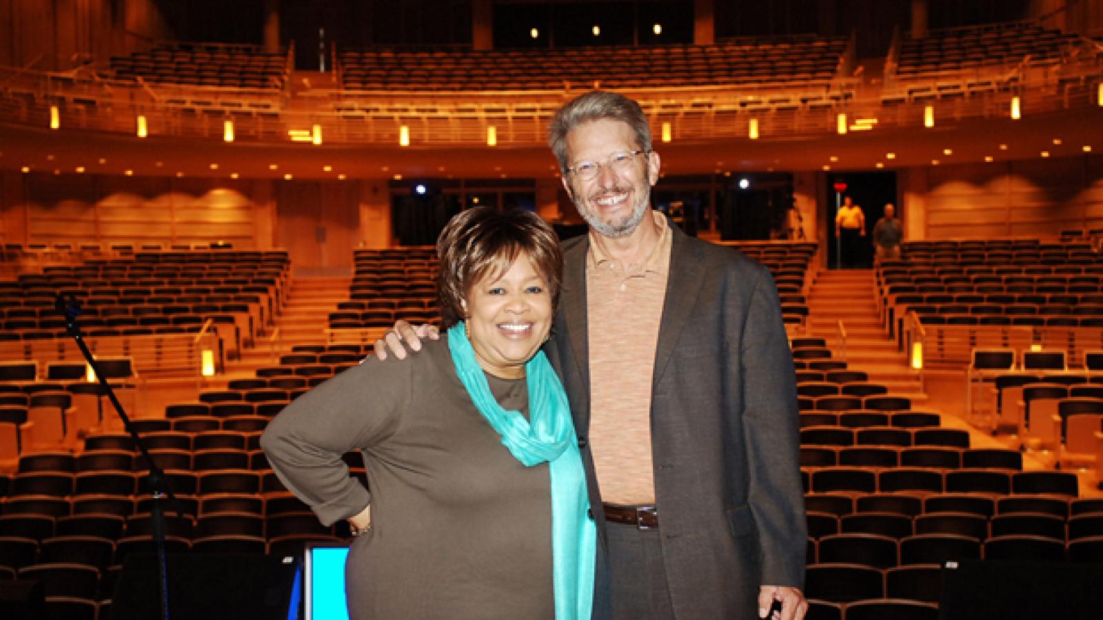 a woman in a brown shirt and a blue scarf stands on a stage with a man with a beard wearing a brown suit.