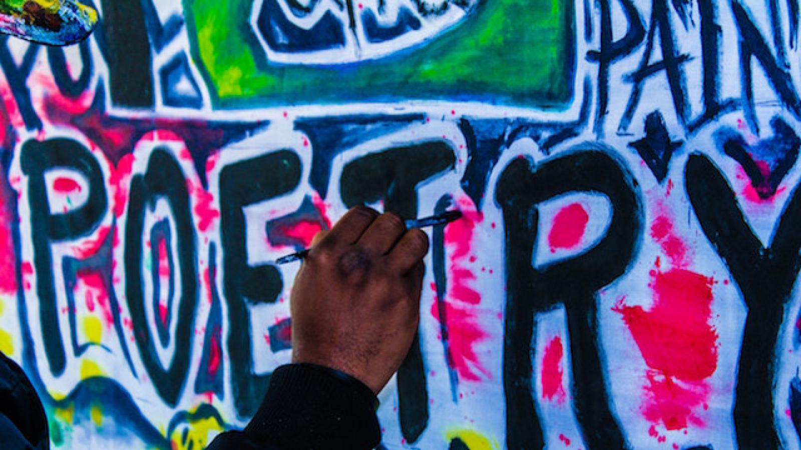 a hand holding a paintbrush working on a wall mural that says poetry