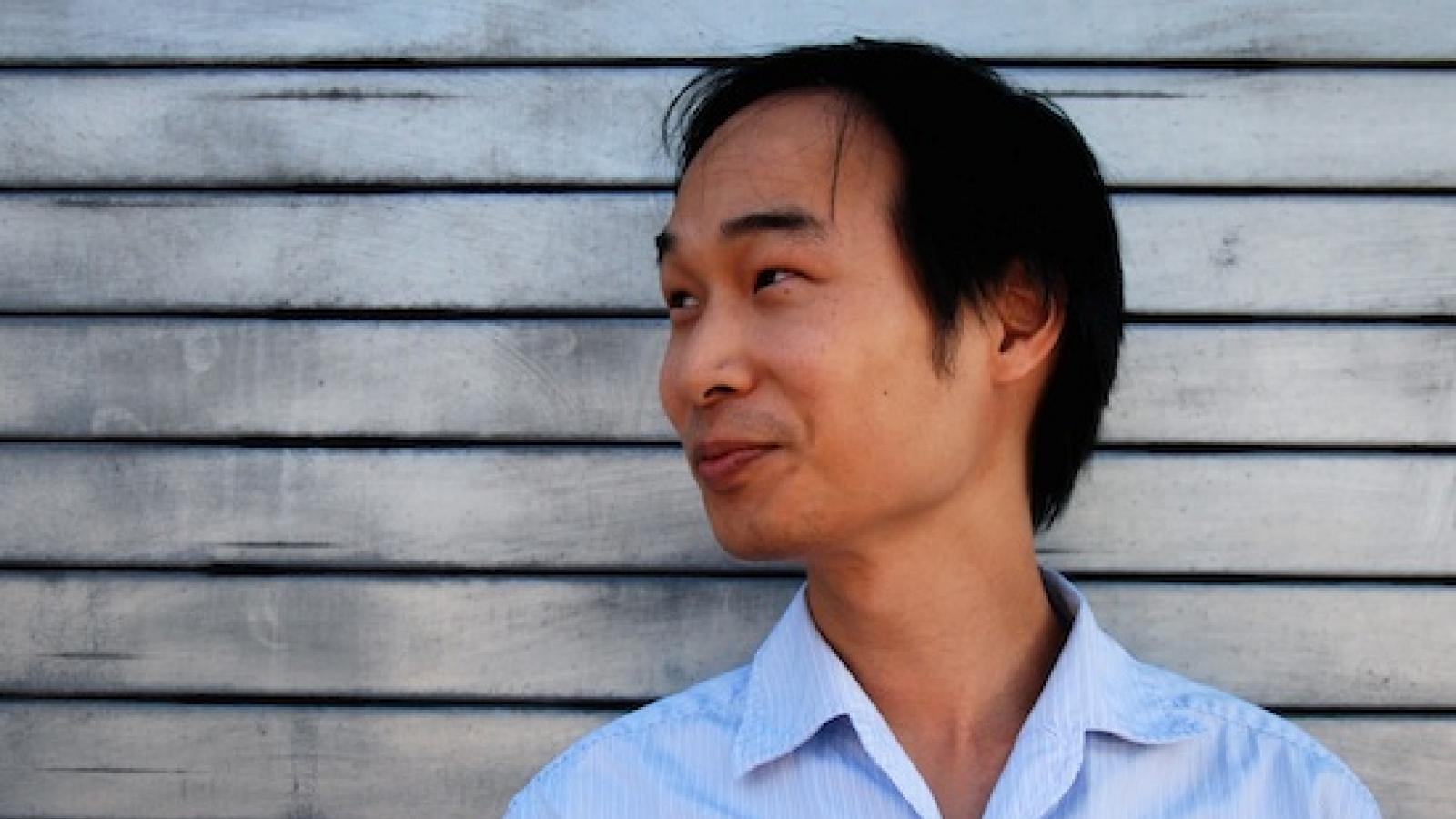 headshot of smiling Asian man in half-profile against background of weathered wood siding
