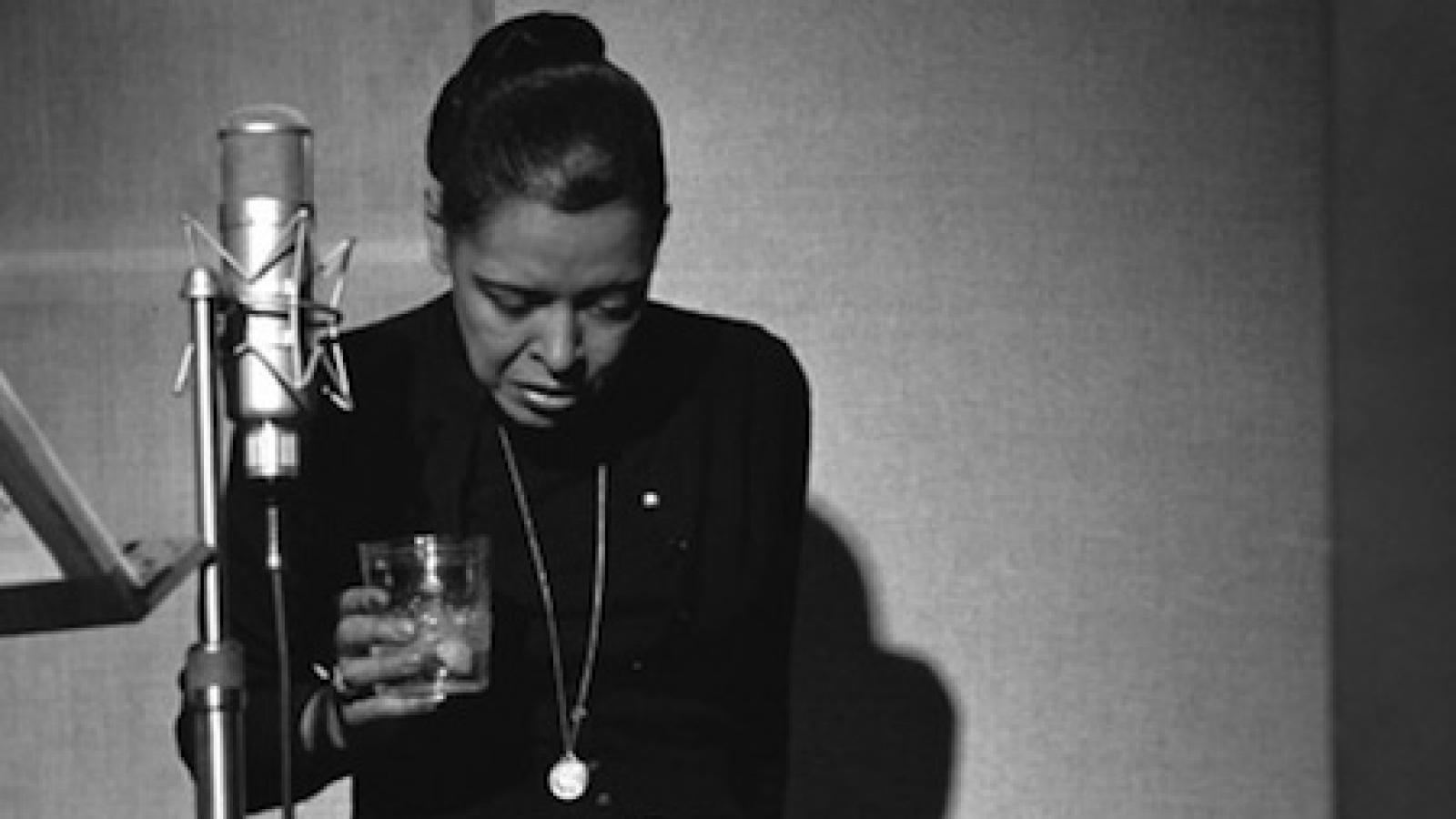 Billie Holiday in black turtleneck stands behind a microphone with her head down and a drink in her hand.