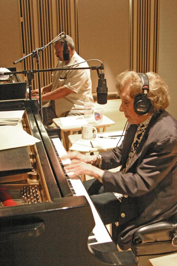 Marian McPartland Woman wearing headphones playing piano with African-American man behind her playing piano.