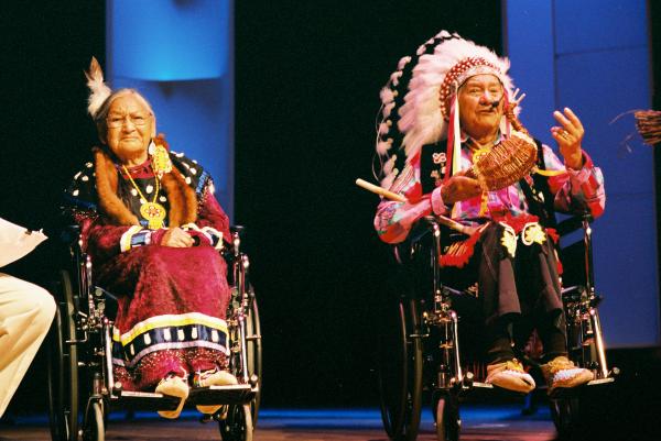 Older Native couple in wheelchairs on stage dressed in traditional Native clothing.