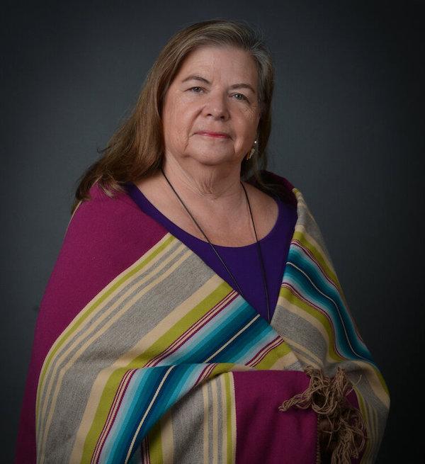 A woman with brown and grey hair is wrapped in a multicolored striped shawl