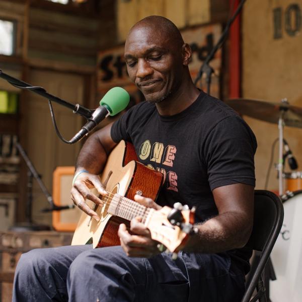 A bald man looks down at his guitar with a microphone in front of him and a drum set behind