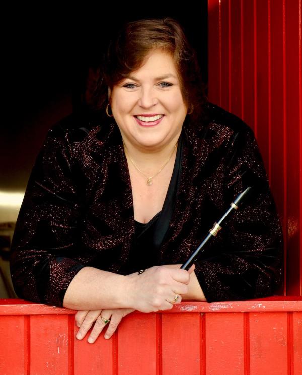 A woman with dark hair holds a black Irish whistle and leans against a latched red wooden half door