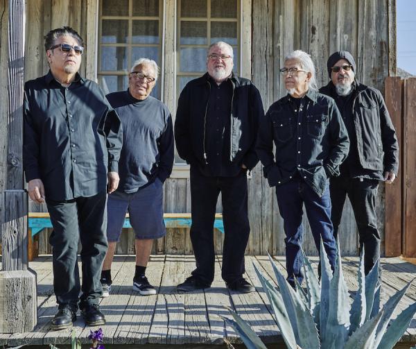 Five men wearing black stand on the porch of a weathered wooden building with a cactus in the foreground