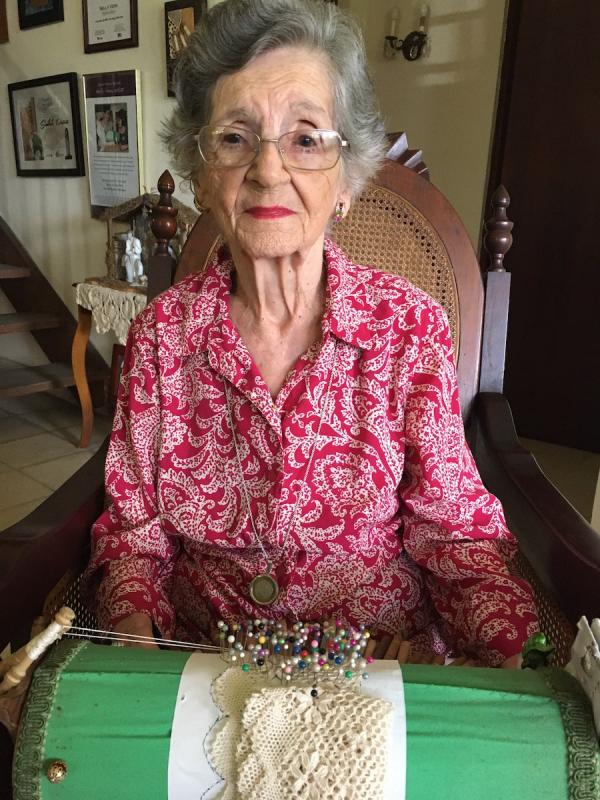 A woman with grey hair and glasses wearing a red and white paisley shirt. In front of her is lace and colorful pins