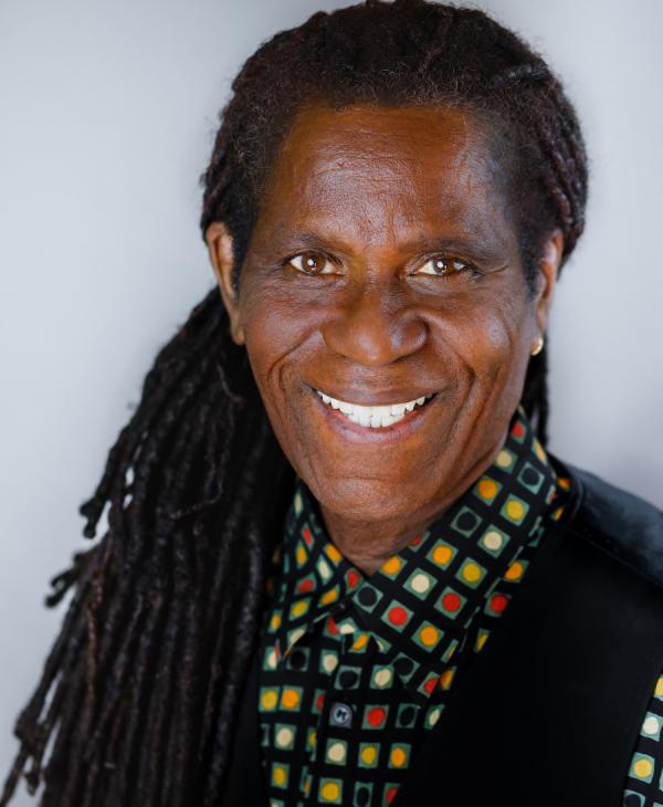 A man with long dark hair wears a colorful shirt and black vest is smiling