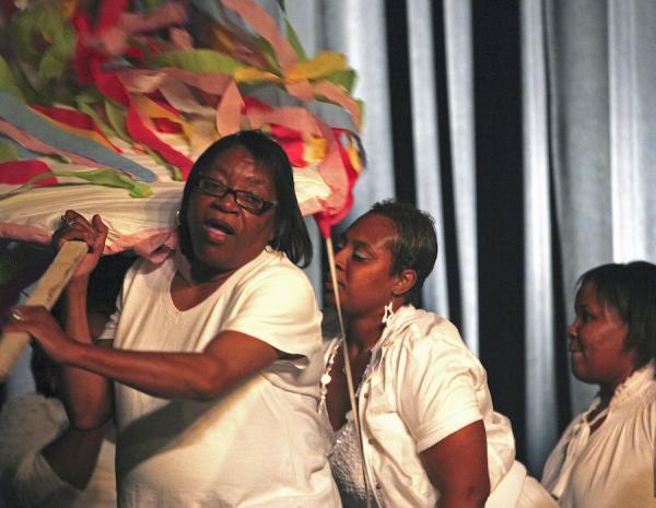 Three women wearing white clothes carry large colorful decorations