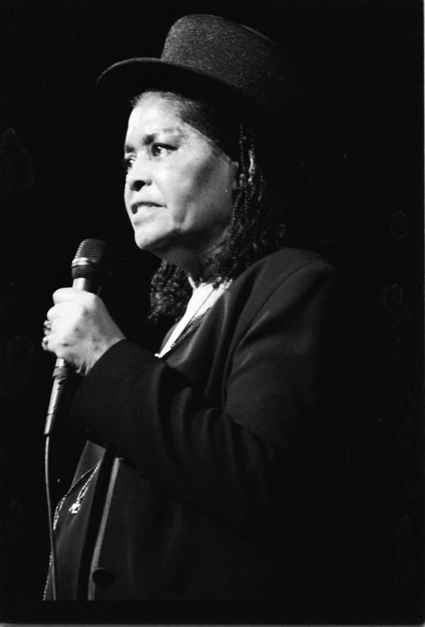 Woman in top hat holding mic on stage. 