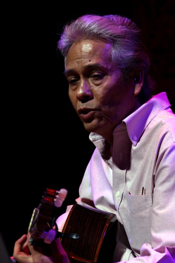 Man in white shirt with white hair brushed back playing guitar on stage.