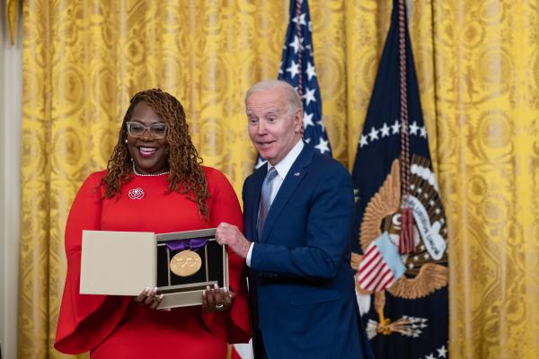 Older white male in blue suit posing with Black woman in red dress in front of flags and gold curtain.