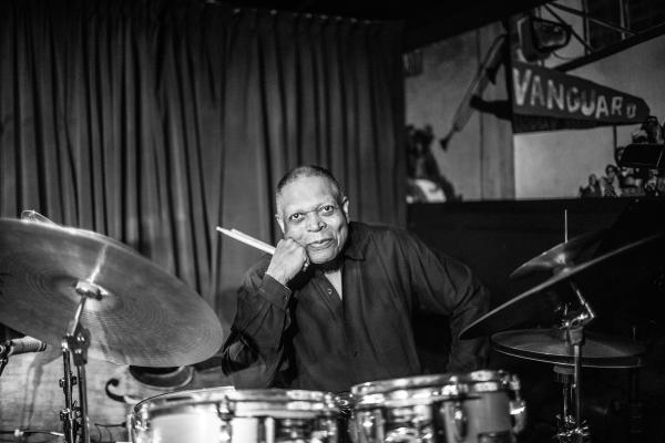 Black and white photo of a man sitting behind a drum kit holding sticks in his right hand. 