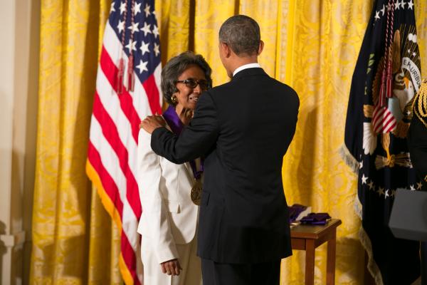 Woman in white suit wearing glasses accepting medal from Black man in dark suit in front of flags and gold curtain