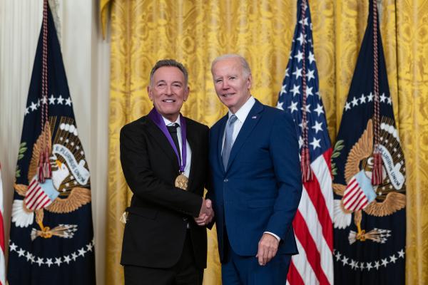 Older white male in blue suit posing with man in black suit in front of flags and gold curtain.