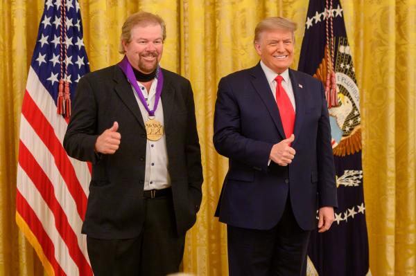 Two men standing in front of US flag holding thumbs up.
