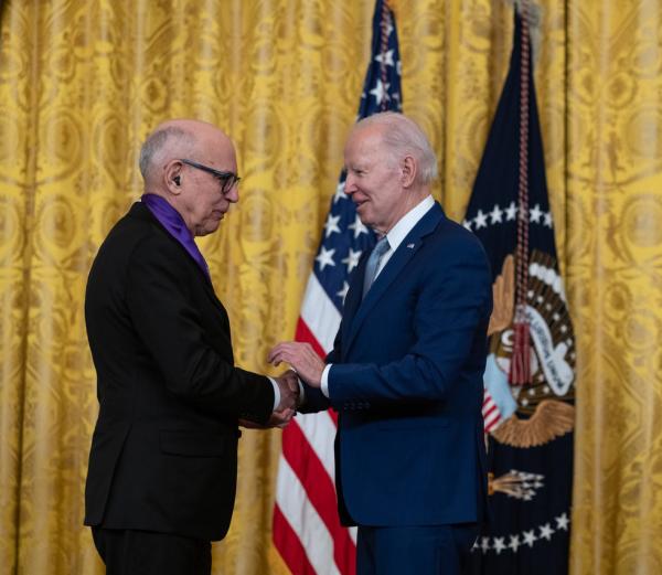 Older white male in blue suit posing with white man wearing glasses in black suit in front of flags and gold curtain.