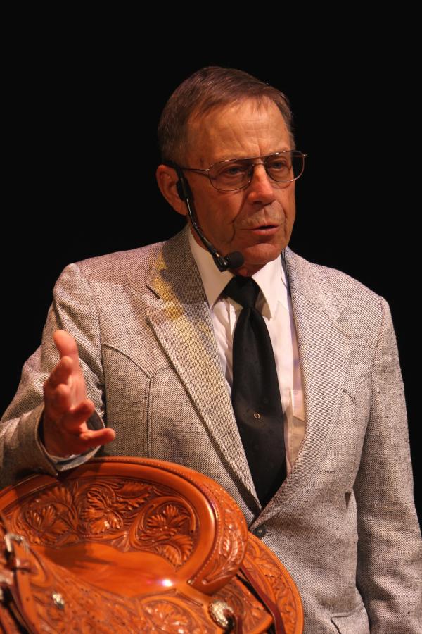 White man in light suit and dark tie wearing glasses with short hair, standing next to a saddle.