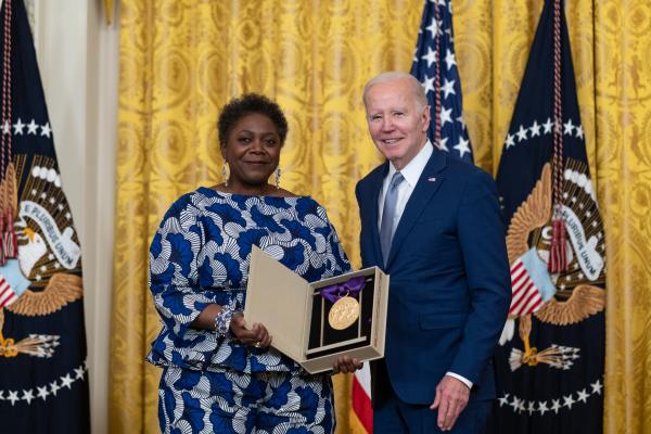 Older white male in blue suit posing with Black woman in blue floral dress in front of flags and gold curtain.