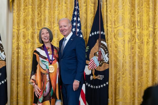 Older white male in blue suit posing with older Asian woman in colorful dress in front of flags and gold curtain.