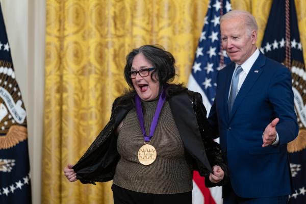 Older white male in blue suit posing with Hispanic woman in glasses wearing a black jacket in front of flags and gold curtain.