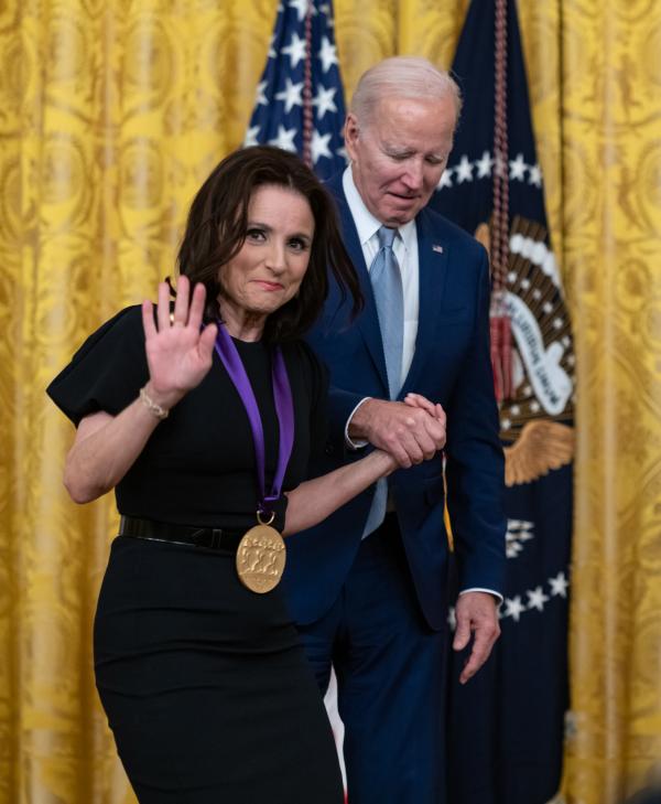 Older white male in blue suit posing with white woman in black dress in front of flags and gold curtain.