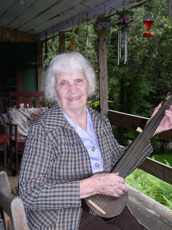 Elderly woman with gray hair sitting on porch playing a banjo.