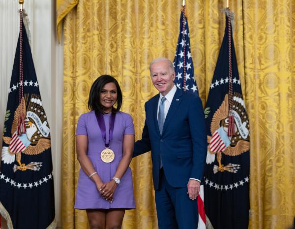 Older white male in blue suit posing with Indian woman in purple dress in front of flags and gold curtain.