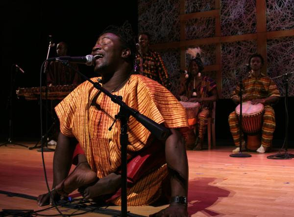 Black man in African shirt and disabled legs singing into a mic on stage. 