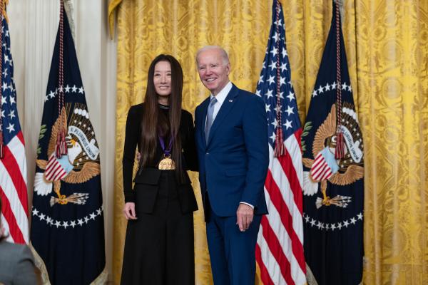 Older white male in blue suit posing with Asian woman in black dress in front of flags and gold curtain.