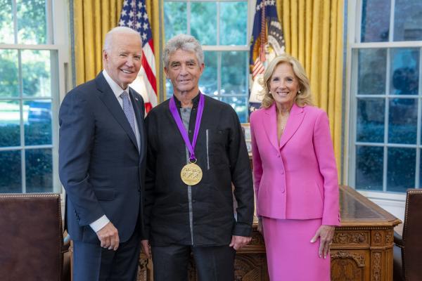 Man with gray hair wearing a medal around his neck surrounded by older man wearing a blue suit and woman with blonde hair wearing a pink suit.