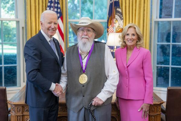 Man with cowboy hat and long gray beard wearing a medal around his neck surrounded by older man wearing a blue suit and woman with blonde hair wearing a pink suit.