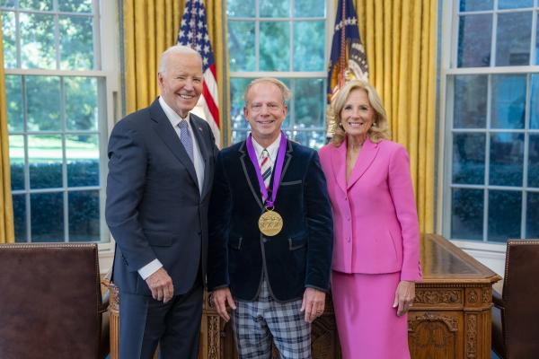 Man in blue velvet jacket wearing a medal around his neck surrounded by older man wearing a blue suit and woman with blonde hair wearing a pink suit.