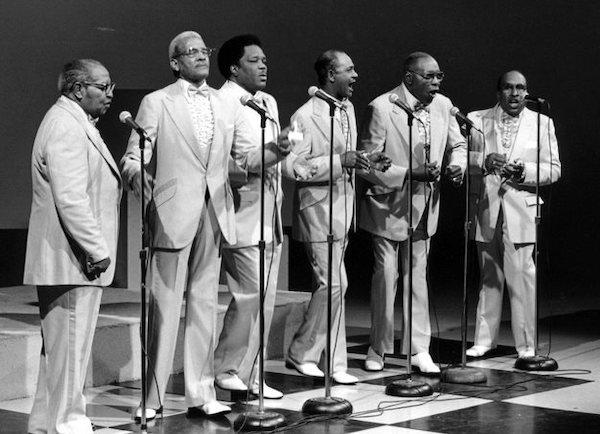 A group of men in suits sing from a satge.