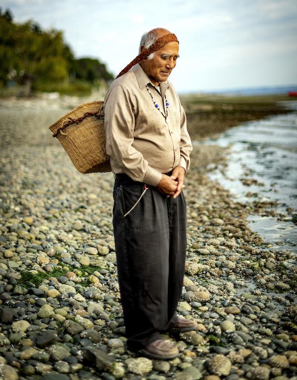A man standing by the water with a basket strapped to his head.