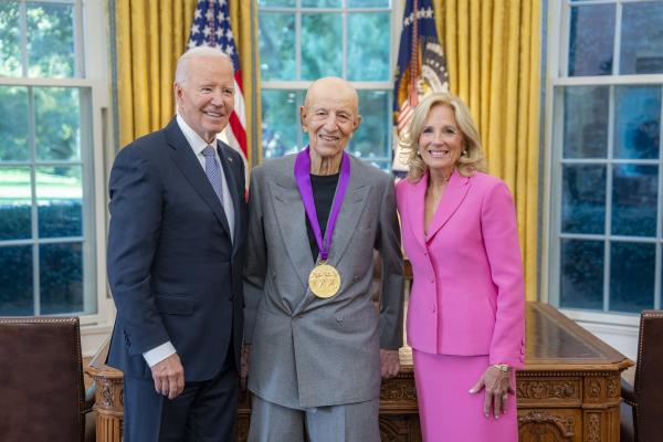 Older bald man in light blue suit wearing a medal around his neck surrounded by older man wearing a blue suit and woman with blonde hair wearing a pink suit.