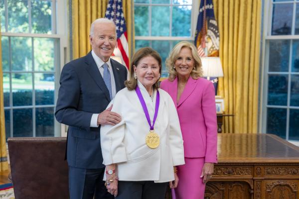 Woman with long brown hair in white shirt wearing a medal around her neck surrounded by older man wearing a blue suit and woman with blonde hair wearing a pink suit.