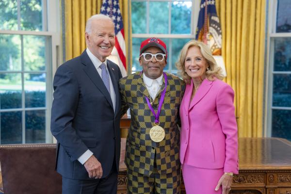 Black man with glasses and New York Yankees cap in a gold and blue checkered suit wearing a medal around his neck surrounded by older man wearing a blue suit and woman with blonde hair wearing a pink suit.