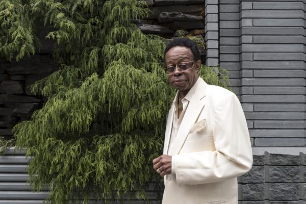 Older Black man in a white suit outside in front of a house with a tree nearby.