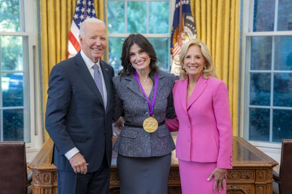 Woman with long dark hair in a gray suit wearing a medal around her neck surrounded by older man wearing a blue suit and woman with blonde hair wearing a pink suit.