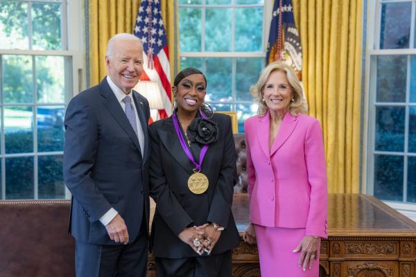 Black woman in dark blue suit wearing a medal around her neck surrounded by older man wearing a blue suit and woman with blonde hair wearing a pink suit.