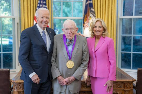 Man in gray suit wearing a medal around his neck surrounded by man wearing a blue suit and woman with blonde hair wearing a pink suit. 